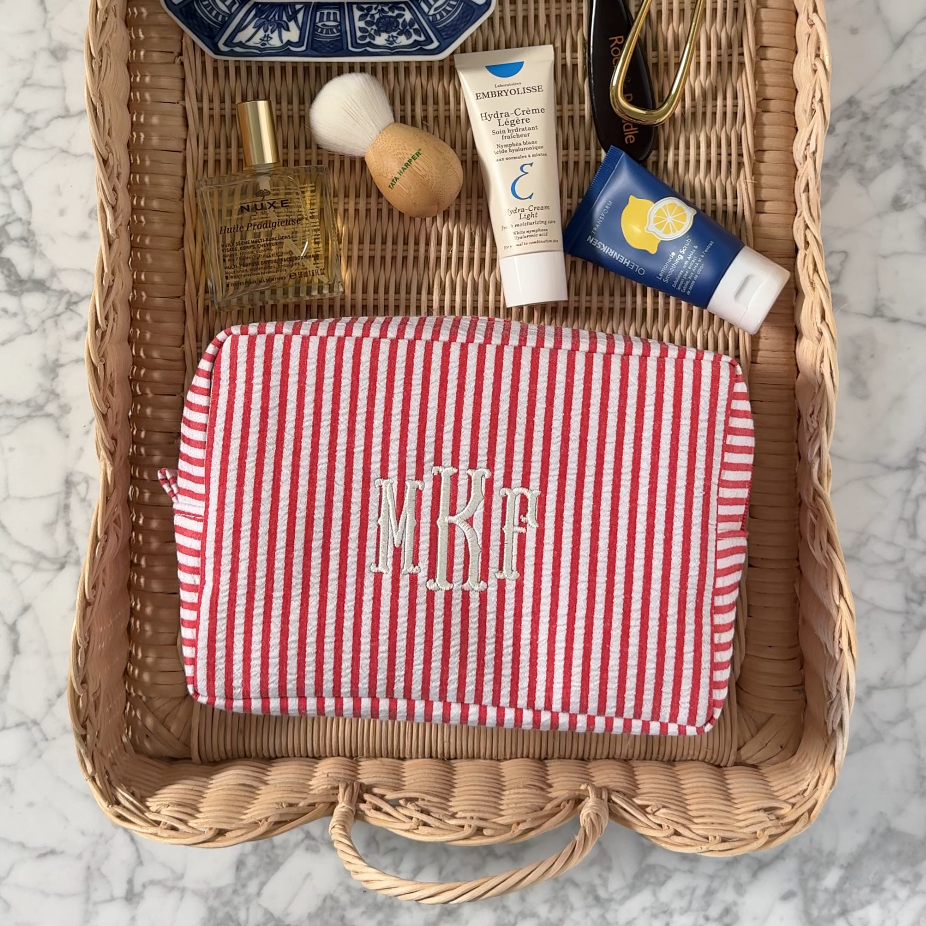 A Casa de Telas red and white striped toiletry bag with an embroidered monogram on a woven tray with skincare products.